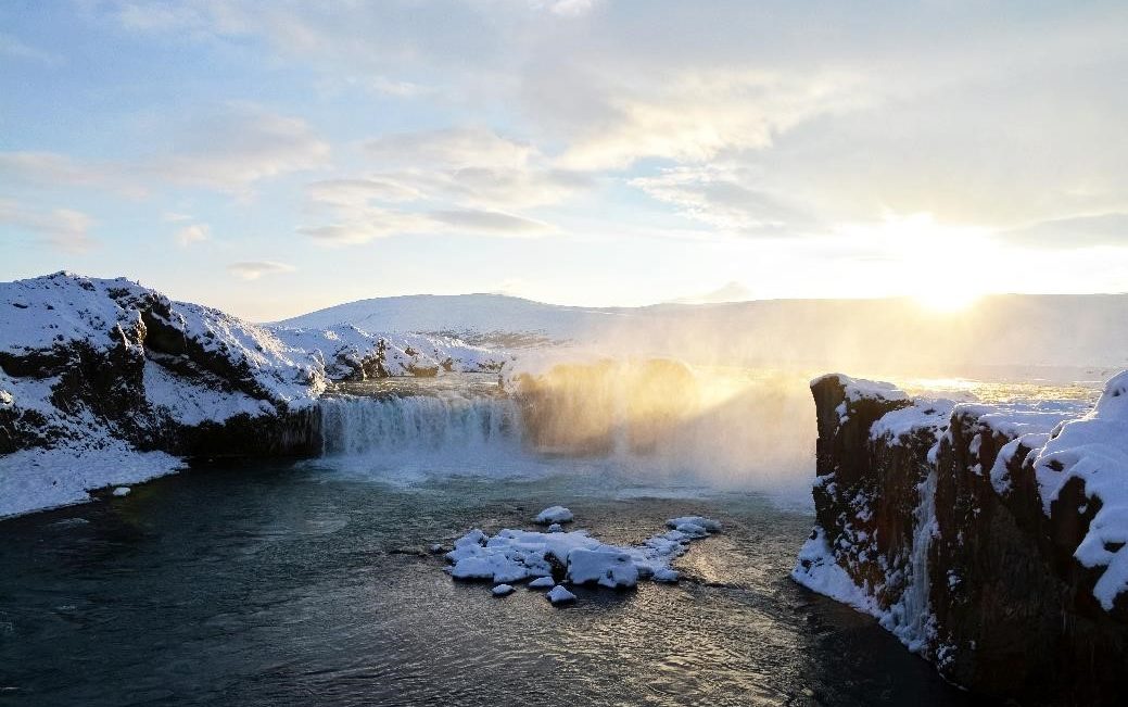 A waterfall in a lake surrounded with snow during sunset.