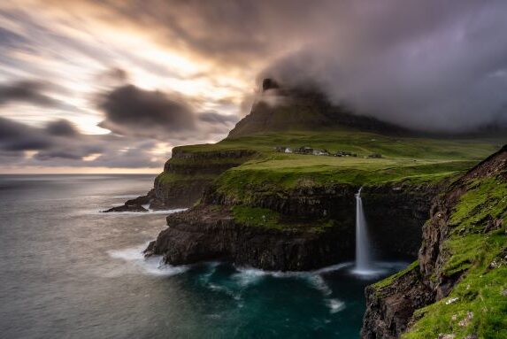 Waterfall into the ocean with clouds covering the top and the sun setting in the background